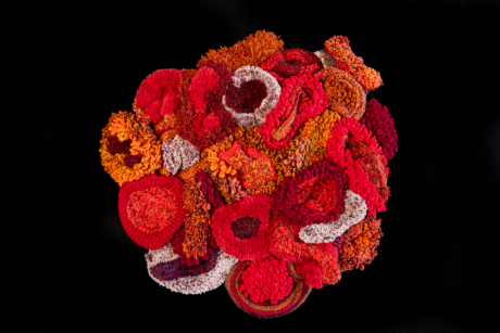 A textured, abstract crochet art piece by Marita Setas Ferro featuring clusters of various sizes and shapes in hues of red and ochre against a black background.