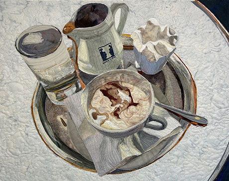 Quilted fabric artwork depicting a coffee cup with whipped cream, spoon, glass of water, creamer pouring coffee, and whipped cream on a tray. Quilted fabric artwork depicting a coffee cup with whipped cream, spoon, glass of water, creamer pouring coffee, and whipped cream on a tray.